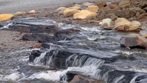 Water-flowing-from-a-wide-stream-flowing-over-rocks-creating-a-small-waterfall-on-beach-in-Kenai-Peninsula-of-Alaska