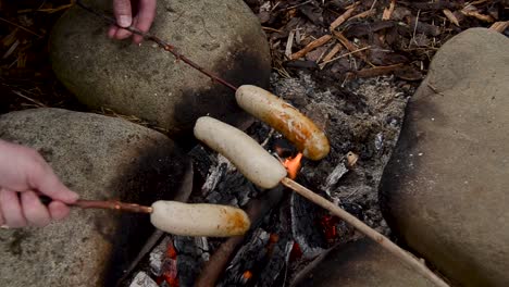 Friends-cooking-sausages-on-a-sticks-for-breakfast-meal-over-flaming-campfire-outdoors