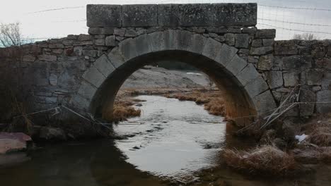 Slow-and-very-low-flight-under-a-roman-times-small-bridge-in-winter,-very-close-to-the-surface-of-the-river-that-goes-under-the-bridge