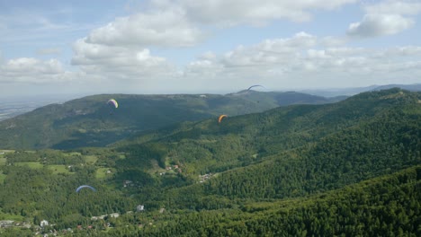 A-group-of-paragliders-fly-above-green-forested-mountains-against-blue-sky-with-white-clouds