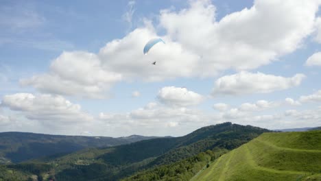 Three-paragliders-flying-over-green-hills,-one-light-blue-paraglider-makes-turn-in-the-center-of-frame-against-a-blue-sky-with-whirling-clouds