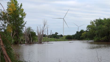 A-flooded-farmers-pond-surrounded-by-trees-with-wind-turbines-in-the-distance-on-a-cloudy-day-in-Nebraska,-USA