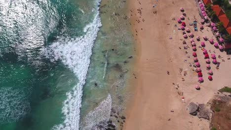 static-drone-shot-of-waves-rolling-in-to-beach-with-pink-umbrellas,-filmed-in-uluwatu,-bali,-indonesia,-tropical-vacation-vibes-beautiful-scene