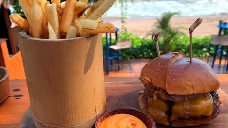 panning-up-view-of-cheeseburger-and-fries-with-sea-view,-filmed-in-seminyak,-bali,-indonesia