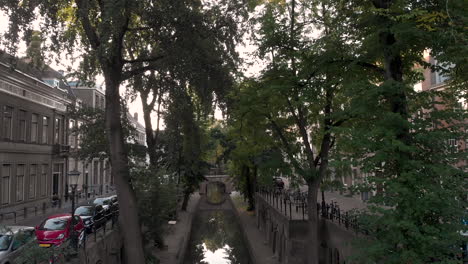 Aerial-tilt-down-showing-early-morning-canal-view-of-the-Nieuwe-Gracht-in-medieval-Dutch-city-of-Utrecht-at-sunrise-between-the-trees-on-the-wharf-water-level