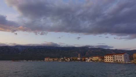 Day-to-night-timelapse-of-small-coastal-town-and-harbor,-mountains-and-rolling-clouds-in-the-background,-colorful-sunset-in-Dalmatian-town-Vinjerac,-Croatia
