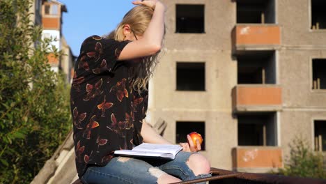 Young-freckled,-blond-,-curly-woman-taking-notes-and-doing-sketches-while-eating-peach