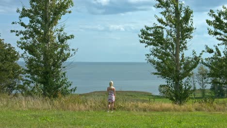 A-woman-standing-on-a-headland,-looking-out-at-the-ocean-and-cloudy-sky,-surrounded-by-greenery