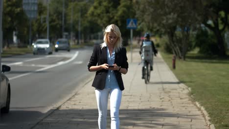 Blonde-beautiful-young-girl-wearing-white-pants,-blue-dress-shirt-and-blacktop-walking-down-a-busy-city-street-with-cars-passing-by-on-her-phone-checking-social-media-and-sending-texts