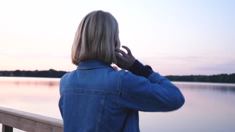 young-short-haired-beautiful-woman-with-jeans-jacked-fixing-her-hair-and-watching-pink-sunset-in-front-of-the-lake