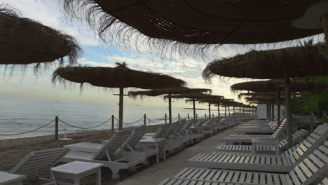 Bewölkter-Strandtag,-Perspektive-Von-Weißen-Sonnenliegen-Und-Strohschirmen,-Marbella-Beach,-Goldene-Meile