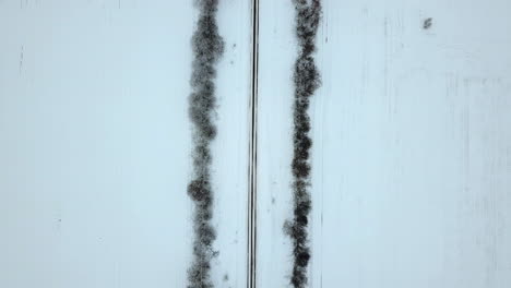 Aerial-footage-of-an-empty,-snow-covered-road-between-birch-alley---TOP-DOWN-view-on-cloudy-Winter-day,-REVEALING-a-slippery-and-dangerous-turn
