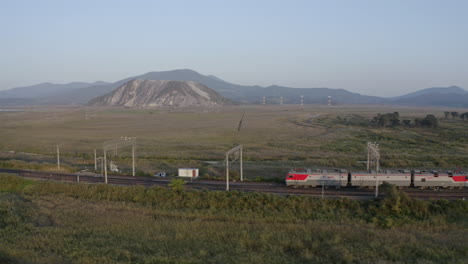 Russische-Eisenbahnzuglokomotiven-Ziehen-Eine-Lange-Leere-Kohlewagenfracht-Durch-Grüne-Felder-Mit-Bergen-Im-Hintergrund-Bei-Sonnenuntergang,-Russische-Föderation