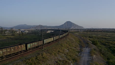 Follow-up-aerial-shot-of-full-loaded-coal-cargo-train-locomotives-pulling-long-carriage-train-approaching-the-intersection-of-the-railways,-on-the-sunset