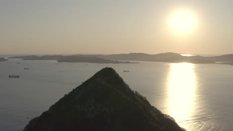 A-summit-of-a-pyramid-shaped-mountain-with-the-bay-and-ships-at-the-anchorage-in-the-background,-and-the-suns-casting-a-big-ray-reflection-on-the-water-surface,-on-the-sunset