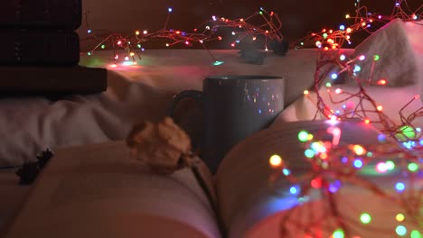 Cozy-background-of-an-open-book-on-a-bed,-next-to-a-mug-and-surrounded-by-colorful-christmas-lights
