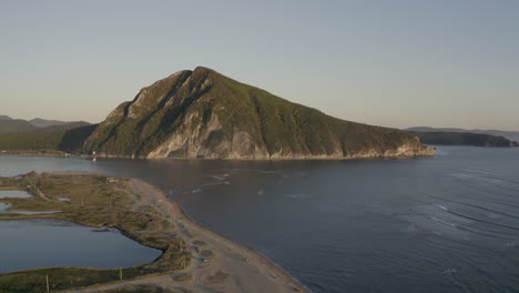 A-landscape-view-of-a-pyramid-shaped-mountain-located-at-a-river-estuary-flowing-into-the-sea,-with-green-vegetation-on-its-sides,-mountain-ridge-in-the-background,-on-the-sunset