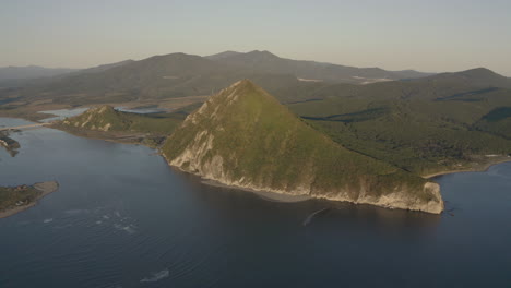 A-landscape-view-of-a-pyramid-shaped-mountain-located-at-a-river-estuary-flowing-into-the-sea,-with-green-vegetation-on-its-sides,-mountain-ridge-in-the-background,-on-the-sunset