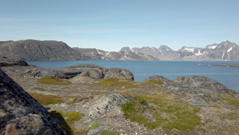 Aerial-view-from-drone-flying-over-rocky-terrain-toward-clear-blue-water-of-fjord-in-Greenland
