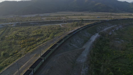 Full-loaded-coal-cargo-carriages-train-stopped-on-a-long-turn-of-a-railway-in-green-fields,-with-road-and-mountain-ridge-in-the-background,-on-the-sunset