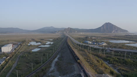 Slow-descent-aerial-shot-of-long-full-loaded-coal-cargo-train-carriage-moving-through-the-fields-with-mountains-in-the-background-on-the-sunset,-Russian-federation