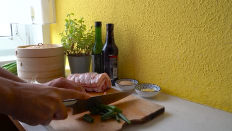 Male-hands-cutting-green-onions-next-to-rolled-up-pork-belly-in-home-kitchen-setting