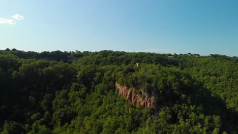 Drone-rising-on-top-of-a-green-valley,-with-summer-woodlands,-some-rocky-mountains-and-a-blue-sky,-Italy,-4k-aerial