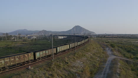 Full-loaded-coal-cargo-freight-train-locomotive-moving-slowly-along-a-high-rise-railway,-in-green-fields-with-mountain-ridge-in-the-far-distance,-on-the-sunset