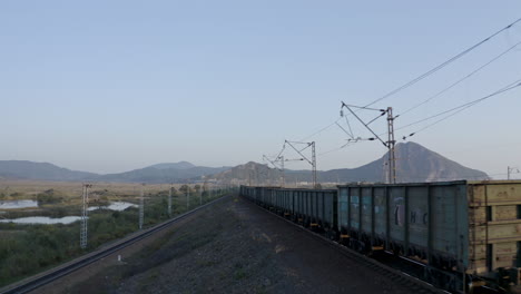 Follow-up-aerial-shot-of-a-long-full-loaded-coal-carriage-train,-passing-through-green-fields,-with-mountain-ridge-in-the-far-distance,-on-the-sunset