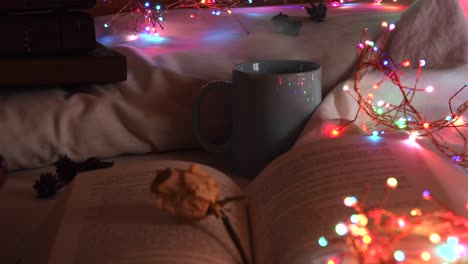 Cozy-background-of-an-open-book-on-a-bed,-next-to-a-mug-and-surrounded-by-colorful-christmas-lights