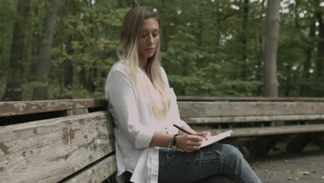 Still-shot-blonde-female-girl-sits-in-the-woods-with-a-note-pad-drawing-taking-notes