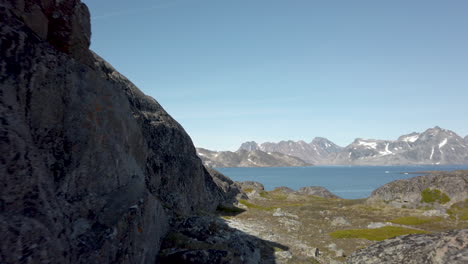 Zu-Fuß-Schmale-Passage-Zwischen-Felswand-Und-Zerklüfteter-Landschaft-In-Richtung-Klares-Blaues-Wasser-Mit-Schneebedeckten-Bergen-In-Der-Ferne-Und-Strahlend-Blauem-Himmel