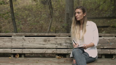 Young-college-aged-woman-sits-in-a-forest-and-draws-sketches-takes-notes-slider-dolly-shot