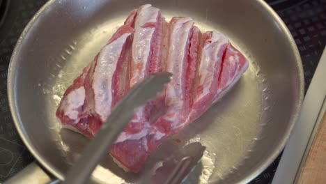Top-down-view-of-cut-of-pork-belly-being-flipped-on-stainless-steel-pan-and-flipped-to-reveal-browning