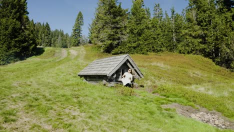 Young-woman-reads-unfolded-map,-looks-for-a-trail,-sitting-in-front-of-small-old-wooden-mountain-hut,-placed-in-the-middle-of-mountain-clearing
