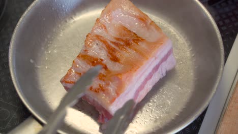 Top-down-view-of-pork-belly-being-cooked-inside-stainless-steel-pan,-moved-around-with-tongs