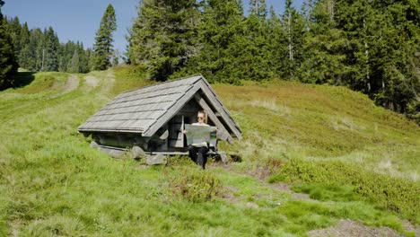 Young-woman-reads-unfolded-map,-sitting-in-front-of-small-old-wooden-mountain-hut,-placed-in-the-middle-of-mountain-clearing