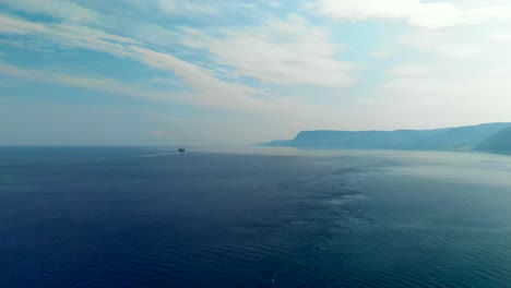AERIAL:-Drone-flying-above-the-Tyrrhenian-Sea,-with-an-incredible-view-of-Calabria's-coastline-in-Italy,-with-a-ferry-sailing-in-the-direction-of-a-mountain