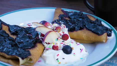 Homemade-crepes-filled-with-cream-cheese,-topped-with-blueberry-jam,-served-with-whipped-cream-and-fresh-fruits-like-apples,-little-strawberries-and-blueberries