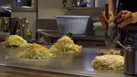 Zubereitung-Traditioneller-Japanischer-Okonomiyaki-Gerichte-Mit-Krautsalat-Und-Fleisch-Auf-Einer-Stahlfläche