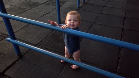 Happy-toddler-boy-stands-alone-on-rubber-playground-mat-leaning-against-blue-jungle-gym-looking-over-bars