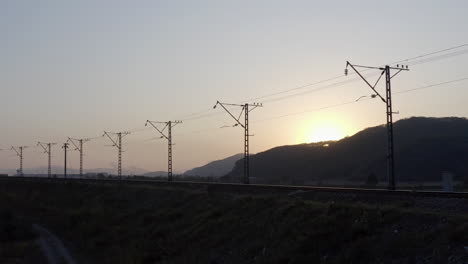 Silhouette-of-empty-railway-with-high-voltage-power-lines-and-sun-hiding-behind-a-hill,-on-the-sunset