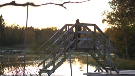 Einsamer-Kleiner-Junge,-Der-In-Der-Abenddämmerung-über-Eine-Kleine-Holzbrücke-Geht,-Stilles-Wasser,-Das-Die-Letzten-Sonnenstrahlen-Reflektiert