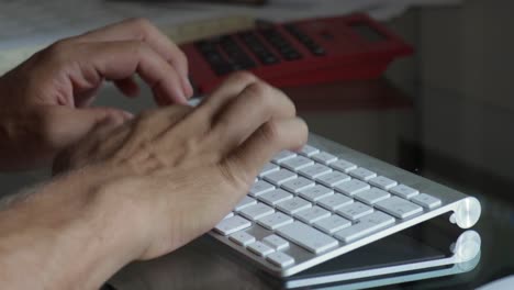 Extreme-Nahaufnahme-Männlicher-Hände,-Die-Auf-Einer-Bluetooth-Tastatur-Auf-Einem-Glastisch-Mit-Taschenrechner-Im-Hintergrund-Tippen
