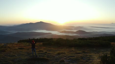 A-hiker-standing-on-a-rock-and-raising-hands-in-a-gesture-of-victory-on-top-of-a-mountain-during-sunrise