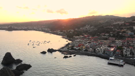 Aerial,-coastal-Italian-town-of-Acitrezza,-sunset-in-background,-arc-right-to-left