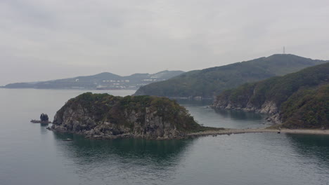 Sea-coastline-with-island-cliffs-with-green-trees-on-the-sides-on-an-overcast-day-with-port-facilities-in-far-distance