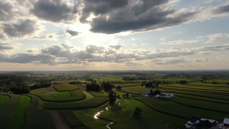 Gorgeous,-cinematic-aerial-shot-of-back-lit-cloud-filled-sky-during-sunset,-beautiful-reflections-on-stream-cutting-through-rural-fields