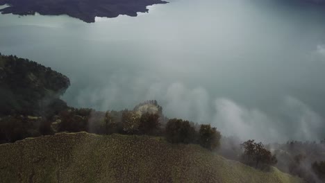 Nubes-Aéreas-De-4k-Que-Se-Vierten-Sobre-El-Borde-Del-Cráter-Del-Monte-Rinjani-Con-El-Lago-En-El-Fondo,-Indonesia