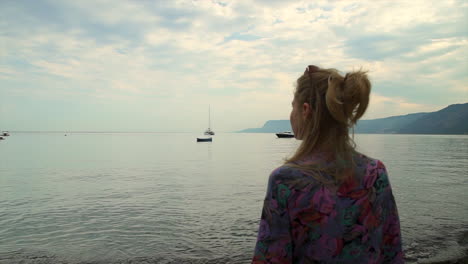Woman-deep-in-thought-on-calm-shoreline-looks-into-distance,-slow-motion-pullback